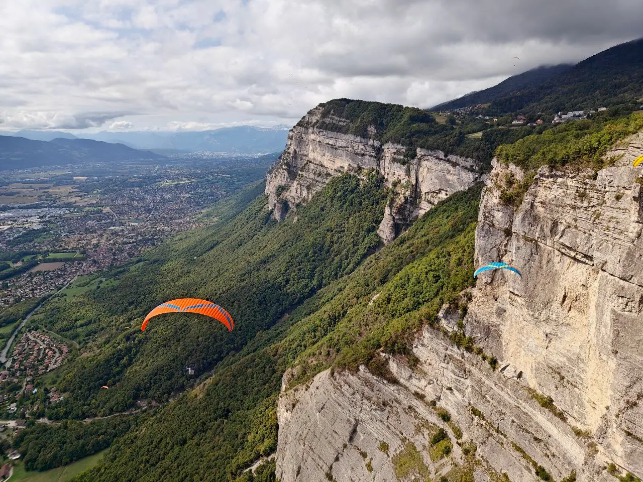 Two paragliders flying close to a tall limestone cliff, with forested slopes falling away to a valley town far below and mountains on the horizon.