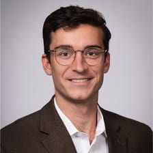 Timothy, smiling, in round wire-rimmed glasses, a brown tweed jacket and an open-collared white shirt.