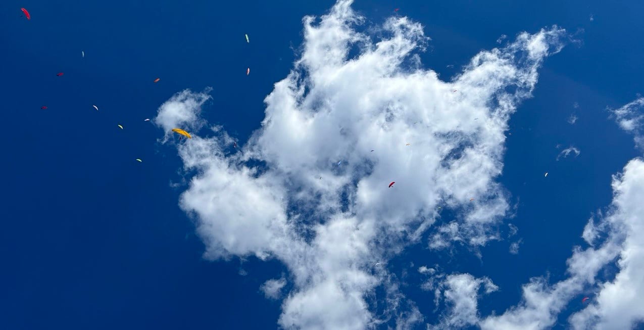 Dozens of paragliders circling together in a thermal beside a tall cumulus cloud, seen from below against a deep blue sky.