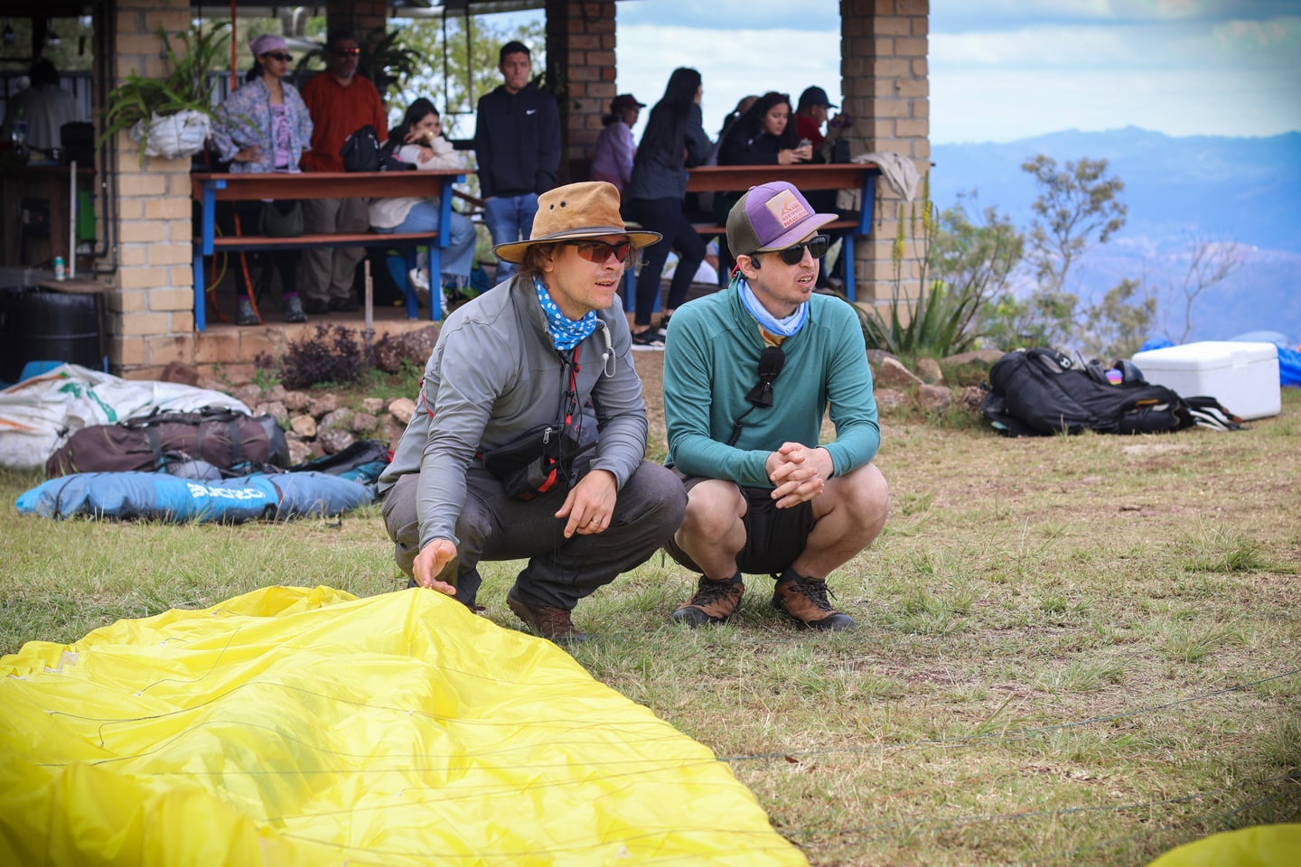Two men crouch on a grassy launch site beside a spread-out yellow paraglider wing, with packed gear around them, a group of people watching from a covered shelter behind, and a hazy mountain valley beyond.
