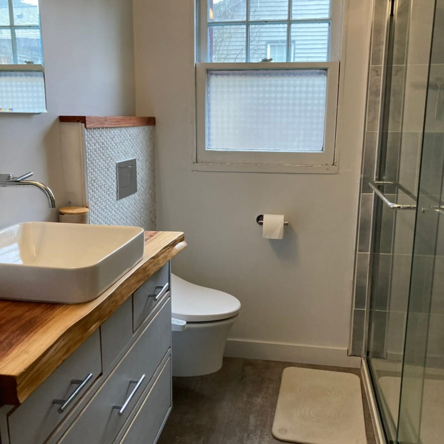 The bathroom after the remodel: a live-edge wood vanity top with a vessel sink and wall-mounted faucet, a tiled glass shower, and new flooring.