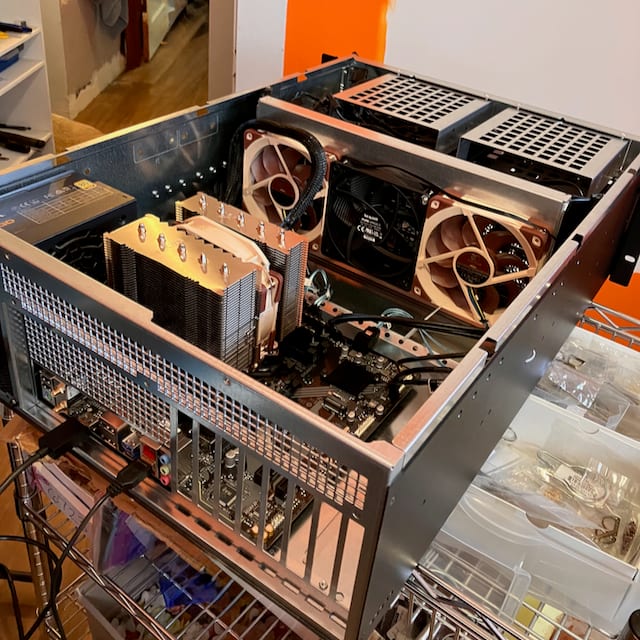 The server mid-build: an open 4U rackmount chassis on a wire shelf, showing the motherboard, a large tower CPU cooler, three case fans, the power supply, empty drive bays, and the PCIe slot covers.