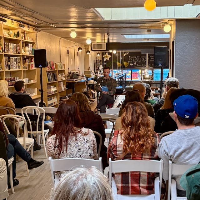 A seated audience, seen from the back of the room, listening to Jonathan read aloud from Unflappable in a small bookshop lined with shelves.